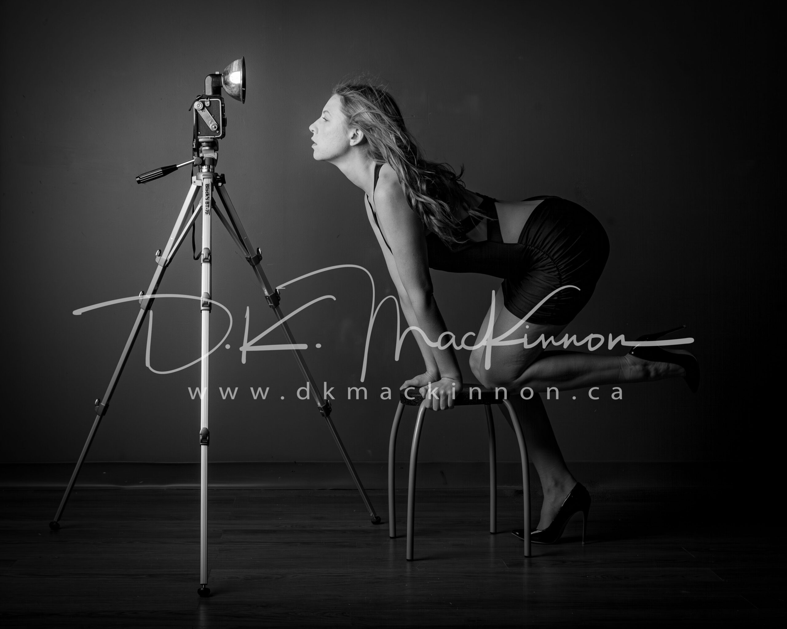 Black and white studio photograph shows a woman in a dark dress leaning toward a vintage camera mounted on a tripod, with a visible photographer's watermark.