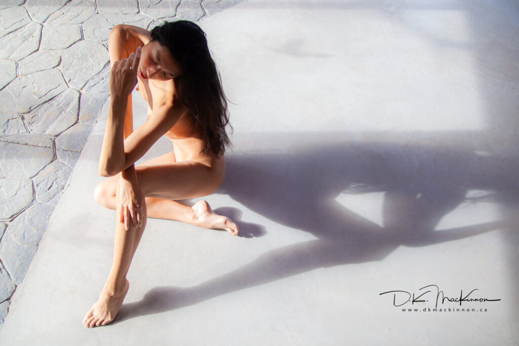 Nude woman with dark hair sits on a gray surface with legs bent, arms raised around head and eyes closed, and dramatic lighting casting long shadows across the surface.