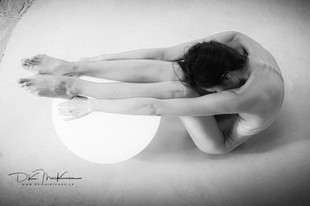 Kalopsia: IMG_3587 Black and white image of a nude woman in a curled pose, cradling her head with her arms while seated on a large, illuminated orb. Her legs are extended in front of her, and her hair is dark and falls over her face. The background is a neutral, textured surface.